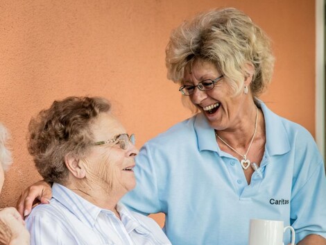 Eine Betreuerin der Caritas und zwei Seniorinnen sitzen lachend auf einer Terrasse. 